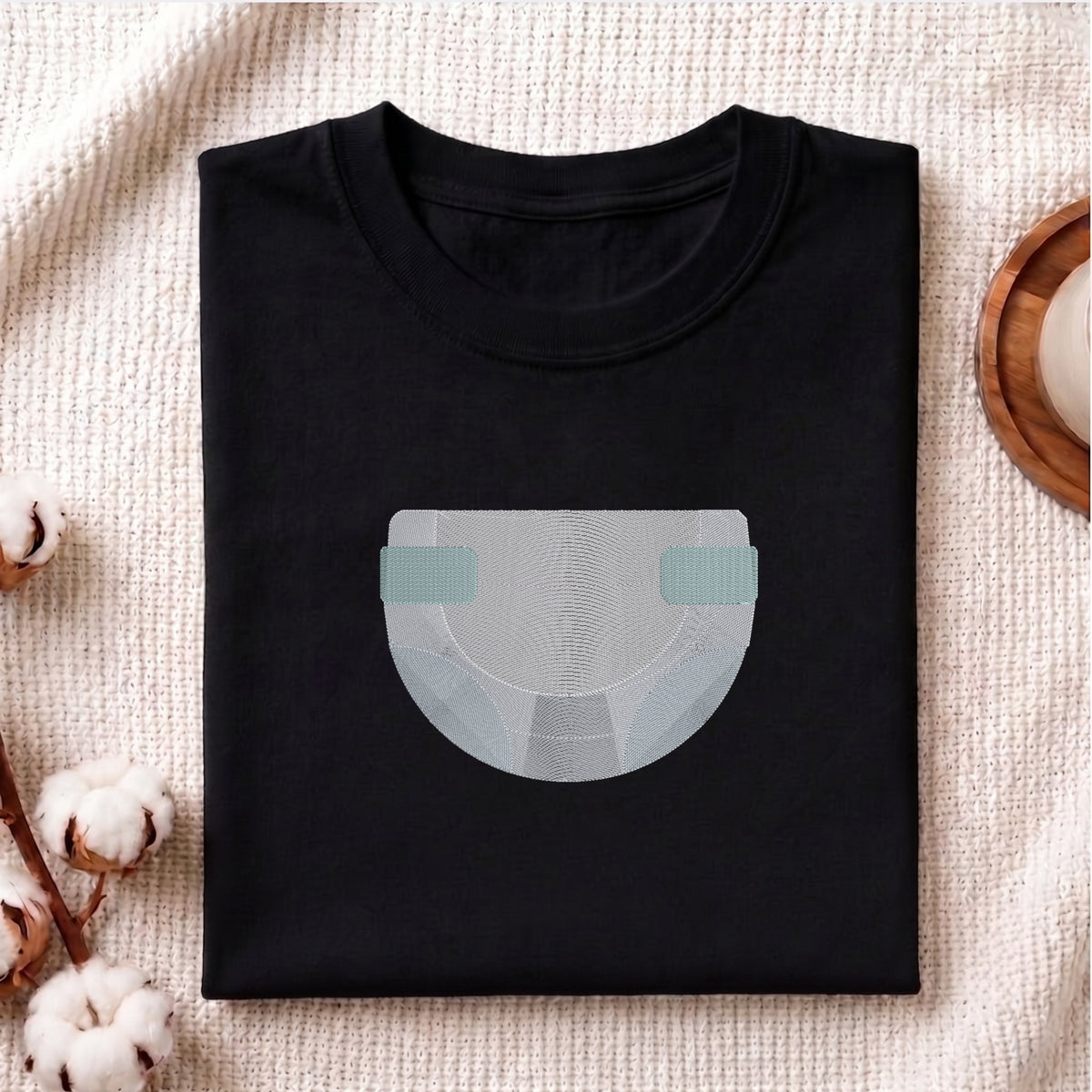 A folded black crewneck t-shirt with a minimalist graphic of a diaper in light gray and mint green, styled on a textured beige blanket with cotton bolls and a wooden coaster.