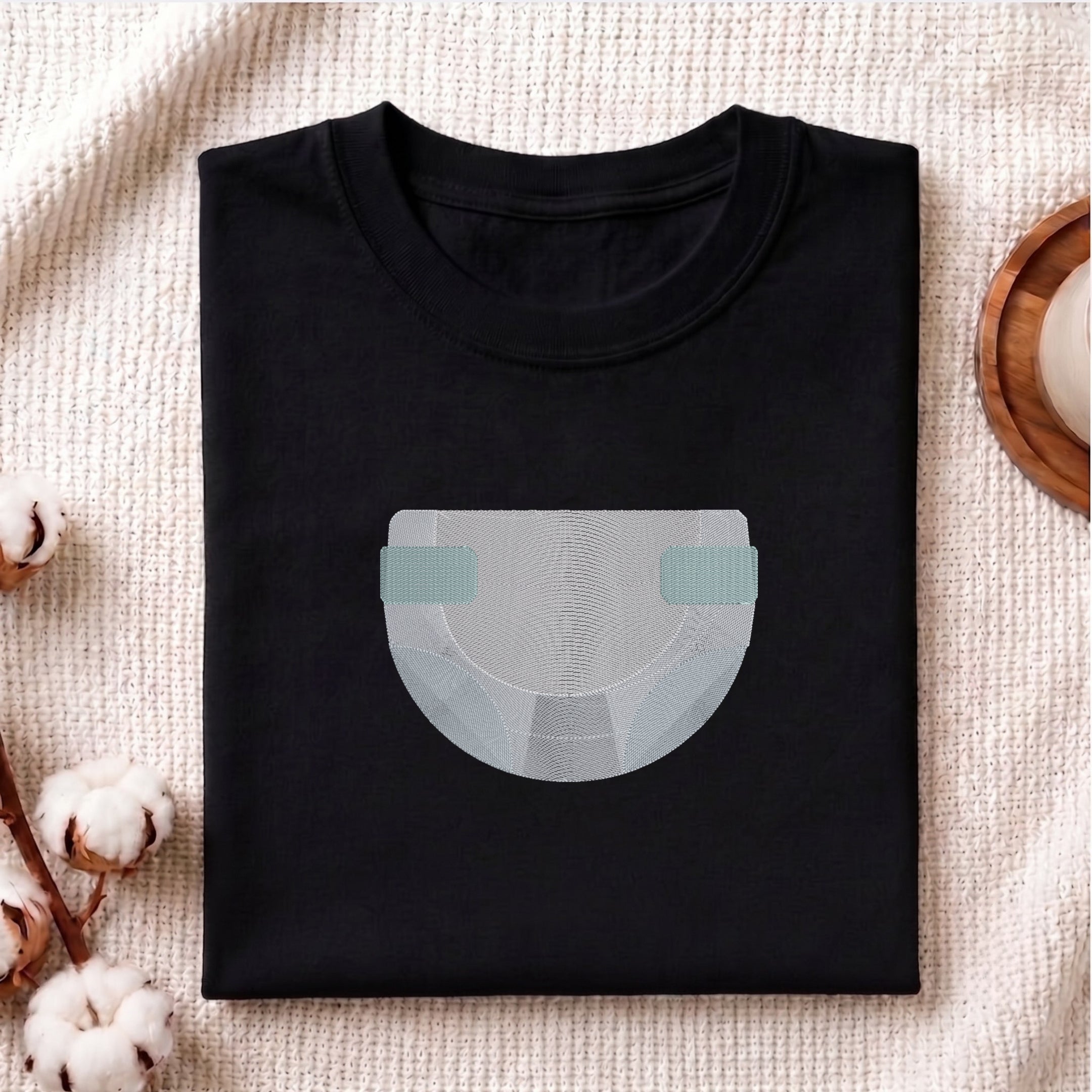 A folded black crewneck t-shirt with a minimalist graphic of a diaper in light gray and mint green, styled on a textured beige blanket with cotton bolls and a wooden coaster.