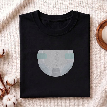 A folded black crewneck t-shirt with a minimalist graphic of a diaper in light gray and mint green, styled on a textured beige blanket with cotton bolls and a wooden coaster.