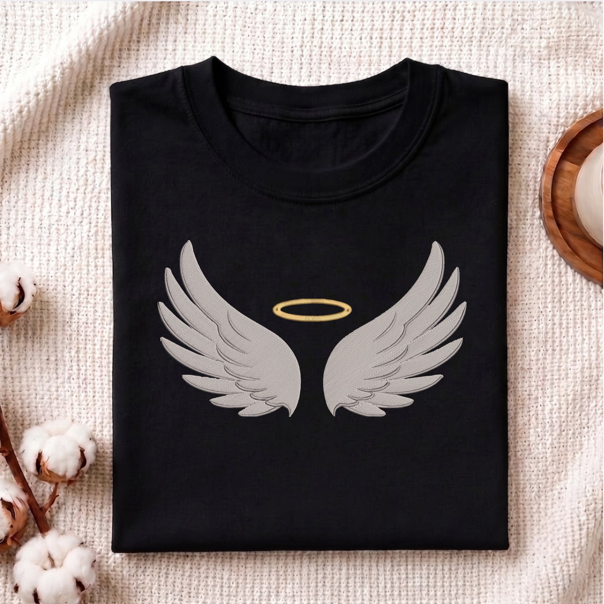 A folded black t-shirt with a graphic of gray angel wings and a gold halo, displayed on a textured beige fabric background with decorative cotton bolls.