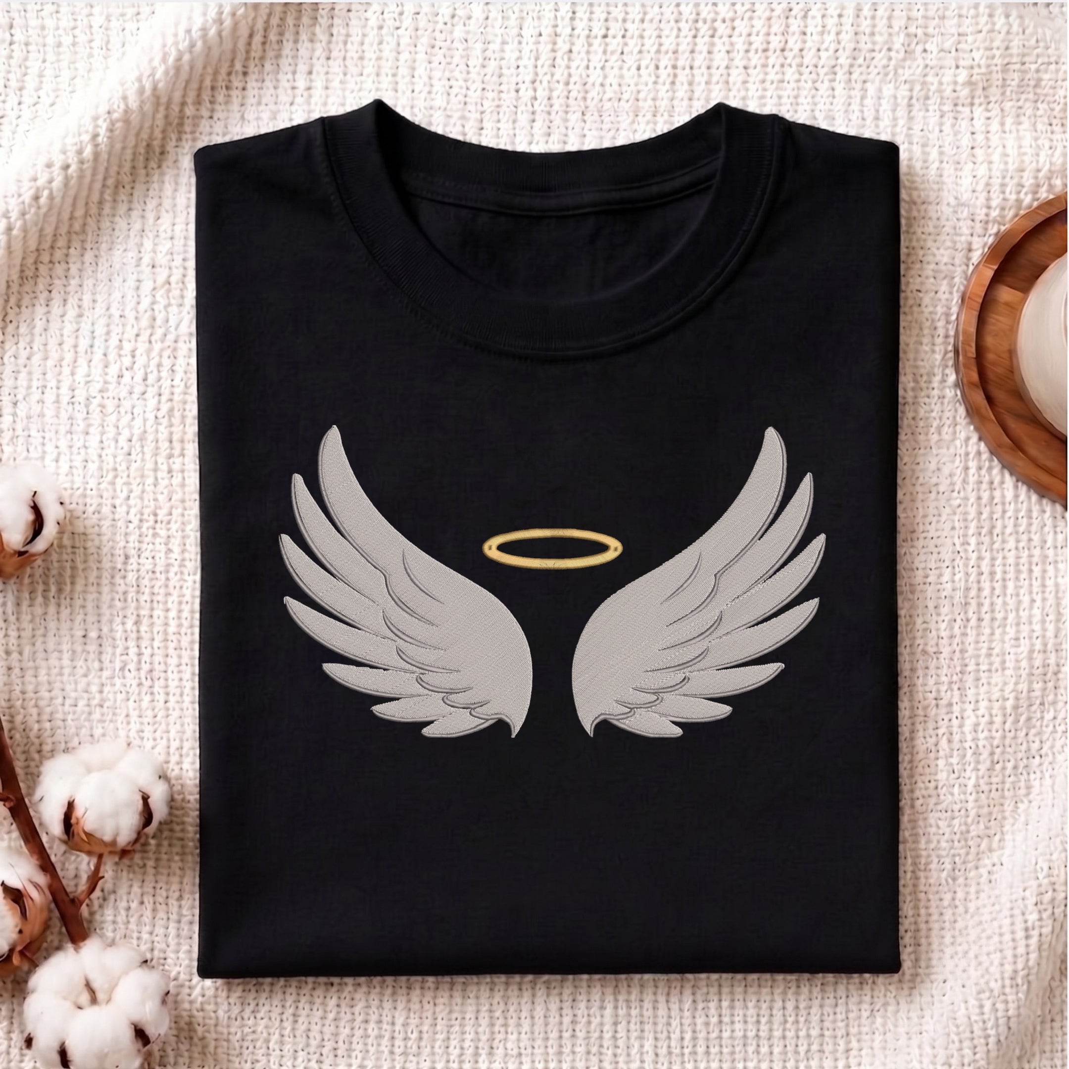 A folded black t-shirt with a graphic of gray angel wings and a gold halo, displayed on a textured beige fabric background with decorative cotton bolls.