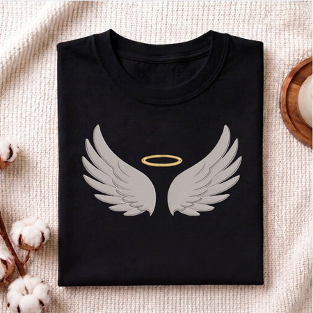A folded black t-shirt with a graphic of gray angel wings and a gold halo, displayed on a textured beige fabric background with decorative cotton bolls.