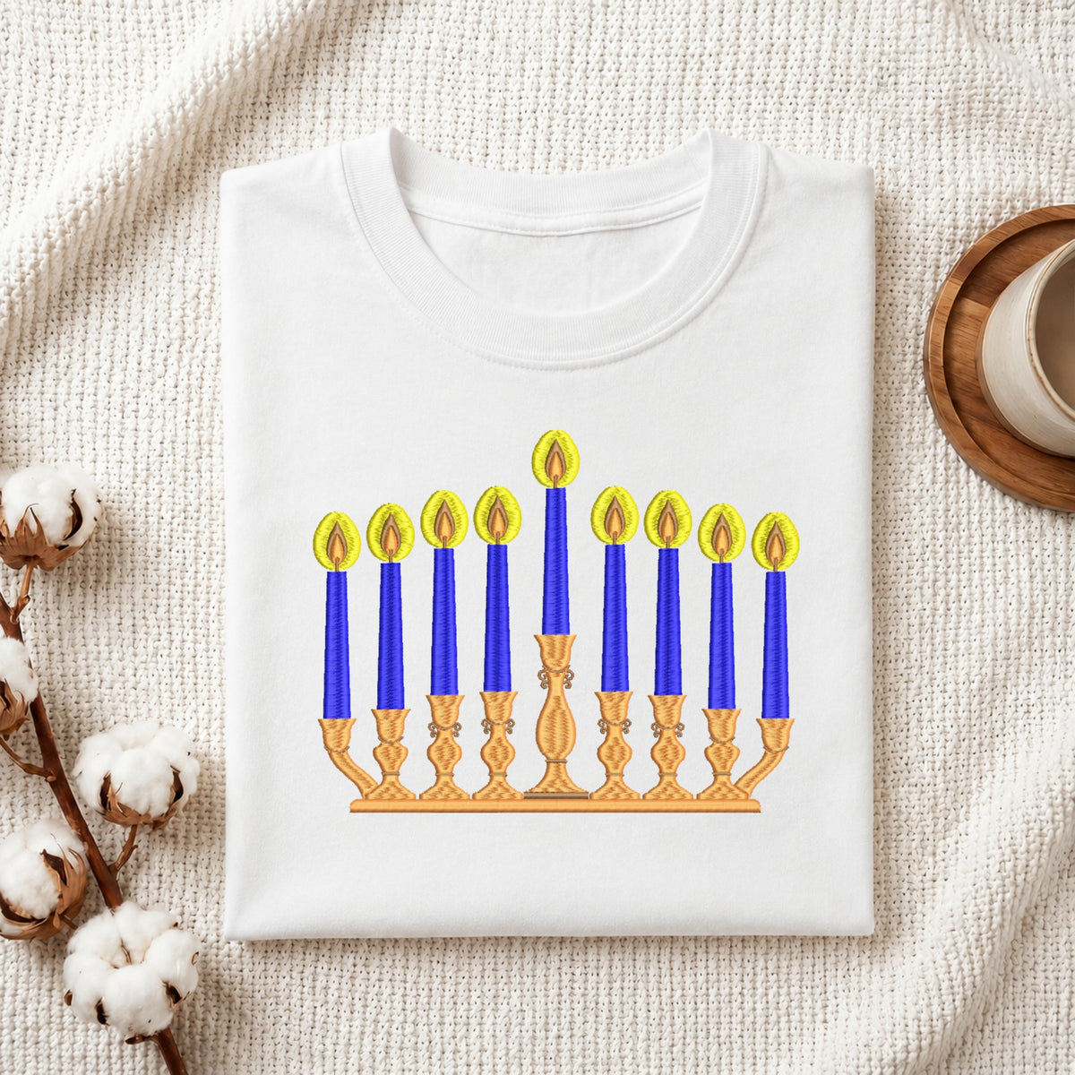 A folded white crewneck t-shirt featuring a printed graphic of a Hanukkah menorah with blue candles and yellow flames, styled on a textured cream fabric with cotton stems and a wooden cup in the background.