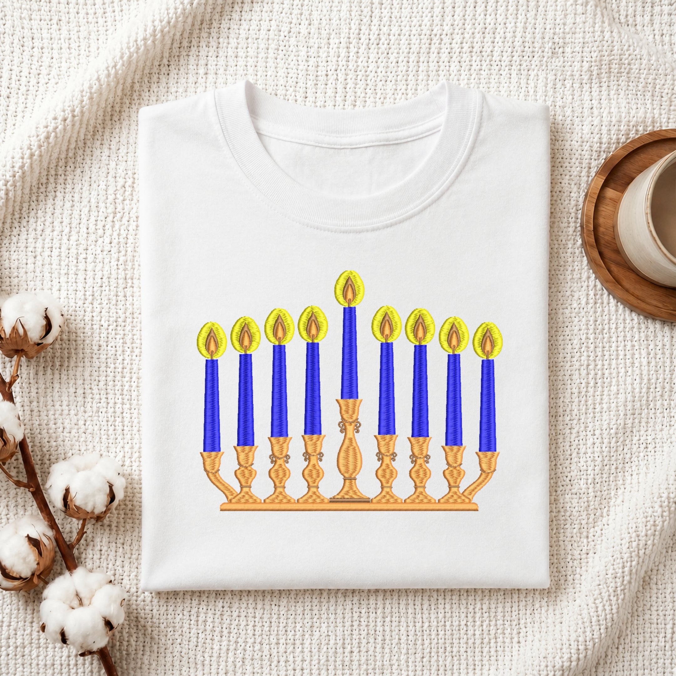 A folded white crewneck t-shirt featuring a printed graphic of a Hanukkah menorah with blue candles and yellow flames, styled on a textured cream fabric with cotton stems and a wooden cup in the background.