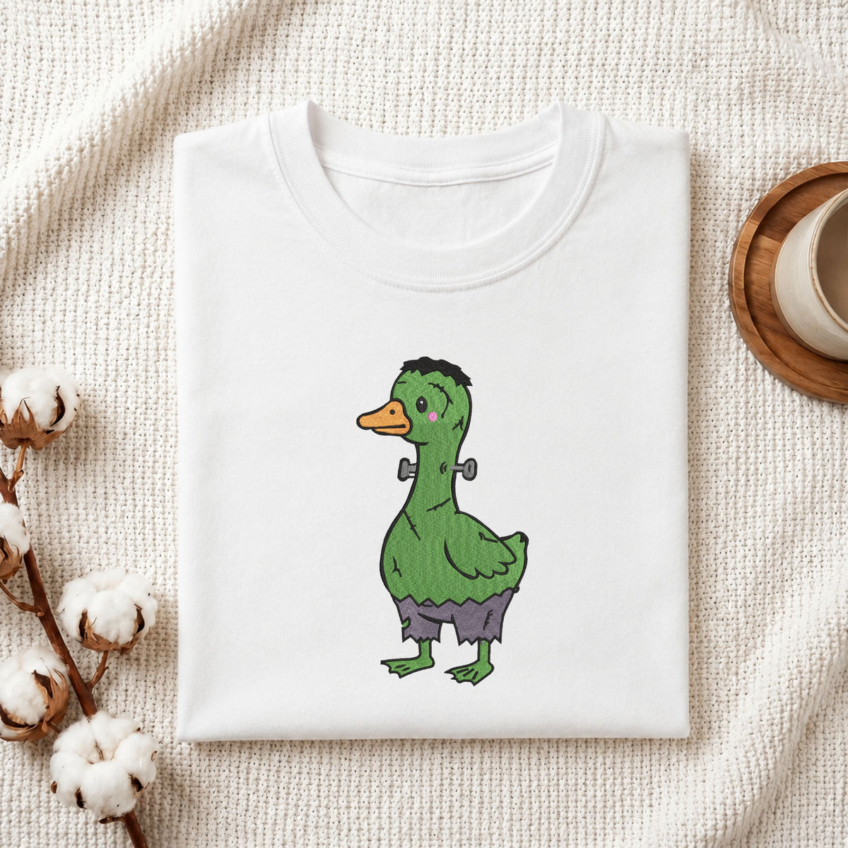 Folded white cotton t-shirt mockup featuring a cute green Frankenstein monster duck Halloween machine embroidery design, styled with cotton stems and a wooden cup