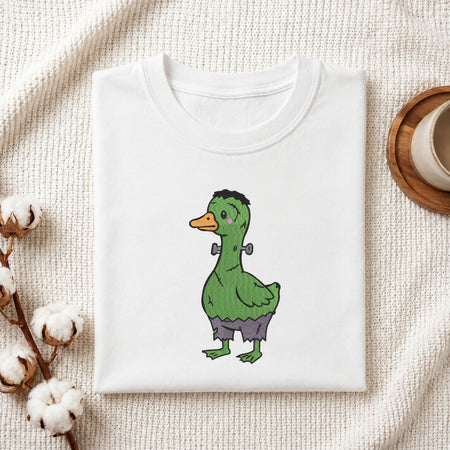 Folded white cotton t-shirt mockup featuring a cute green Frankenstein monster duck Halloween machine embroidery design, styled with cotton stems and a wooden cup