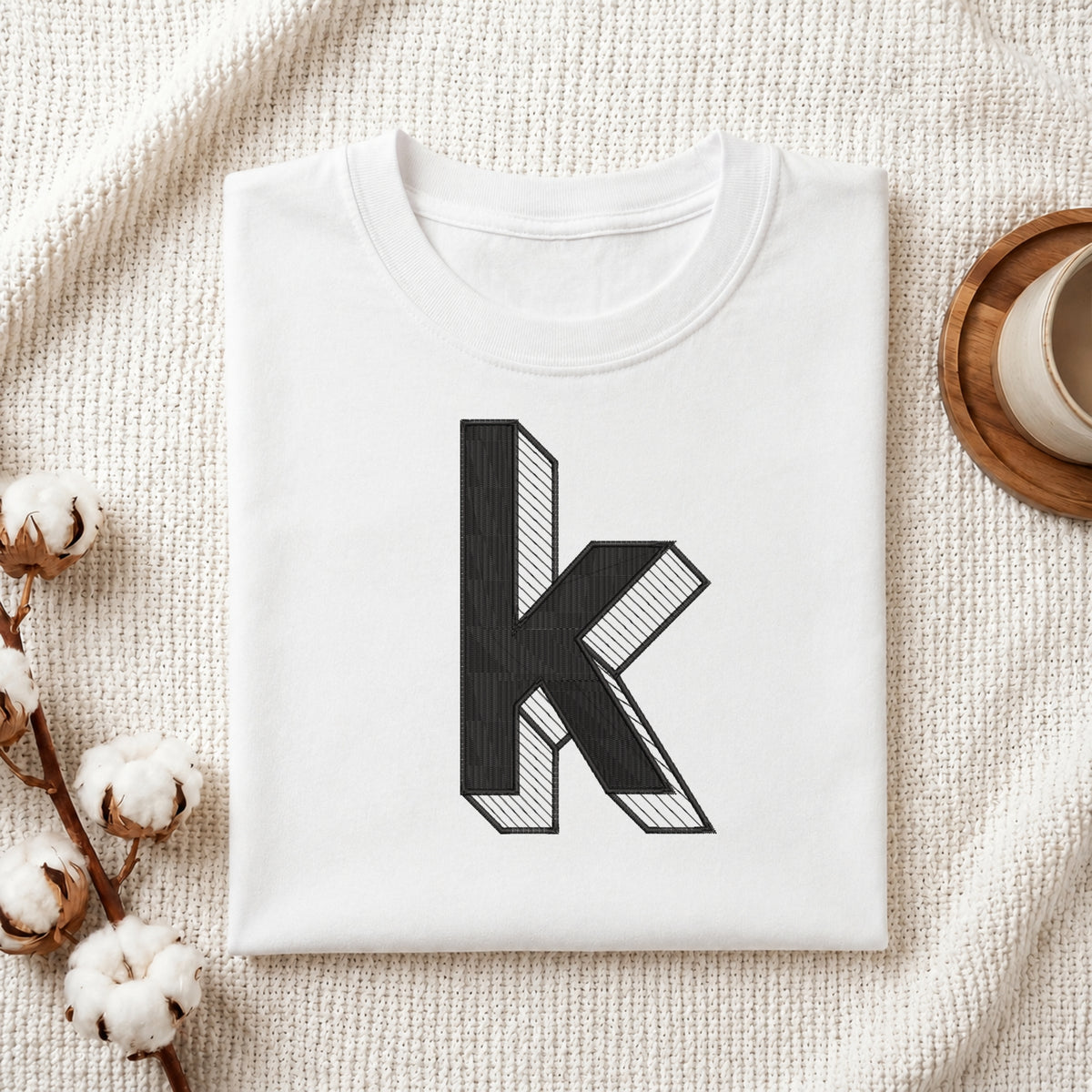 A folded white t-shirt with a large black 3D-effect letter 'k' printed on the front, styled on a textured cream blanket with cotton bolls and a cup of coffee in the background.
