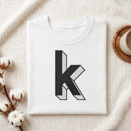 A folded white t-shirt with a large black 3D-effect letter 'k' printed on the front, styled on a textured cream blanket with cotton bolls and a cup of coffee in the background.