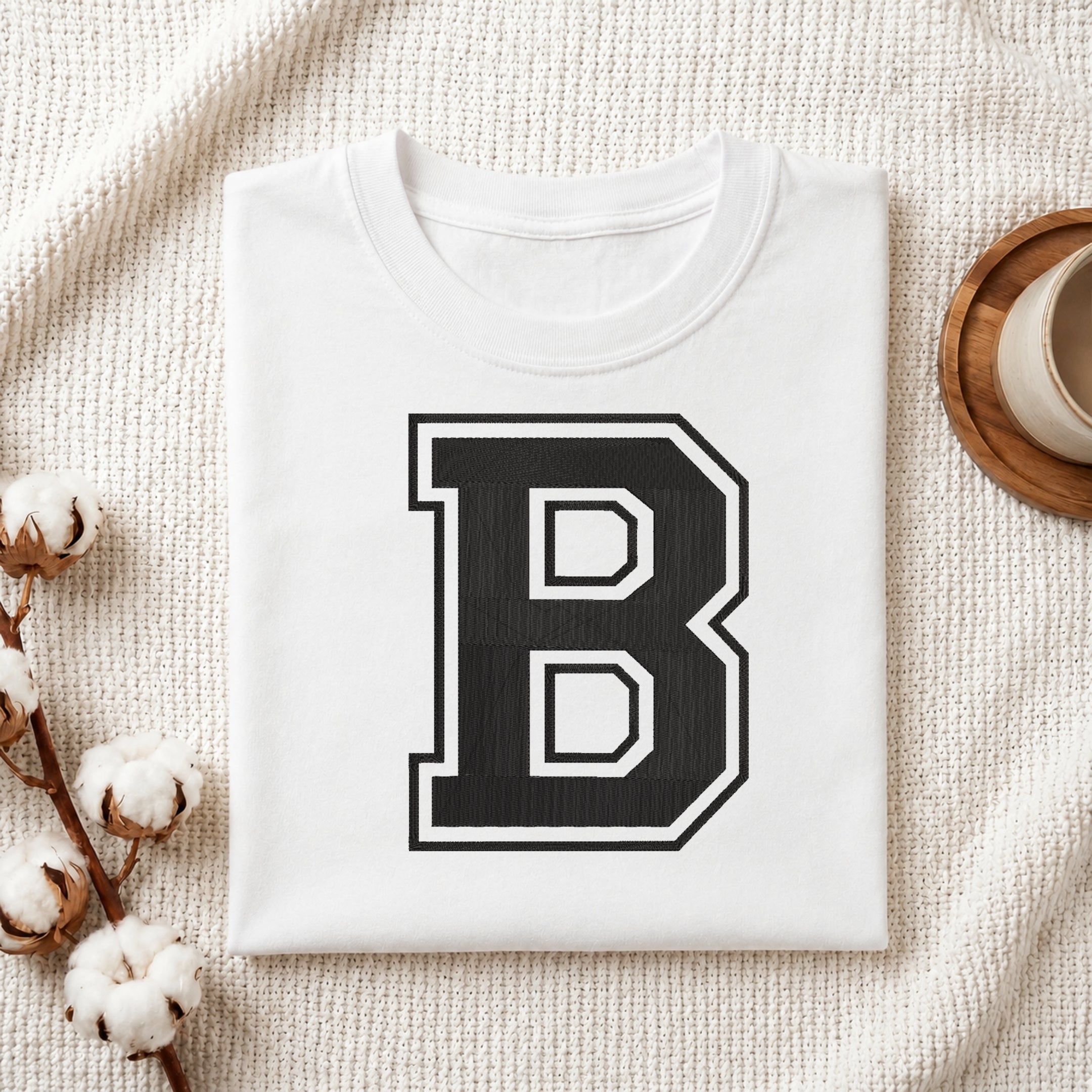 A folded white T-shirt with a large black block letter B outlined in white on the front, styled on a textured cream fabric with cotton stems and a small cup in the corners.