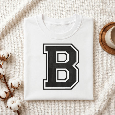 A folded white T-shirt with a large black block letter B outlined in white on the front, styled on a textured cream fabric with cotton stems and a small cup in the corners.