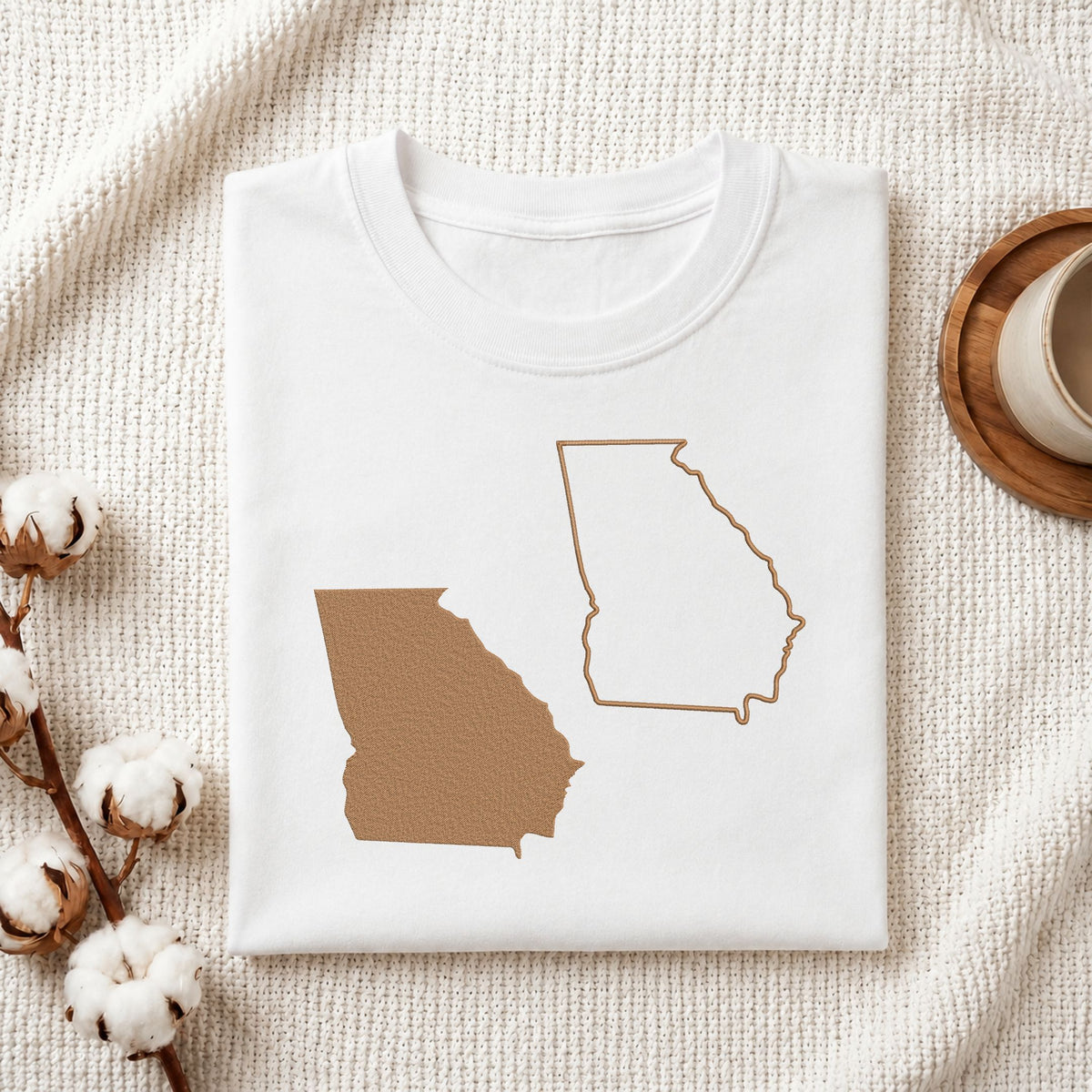 A folded white T-shirt with a graphic of the state of Georgia in tan, styled on a textured cream fabric with cotton stems and a cup on a wooden coaster.