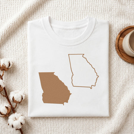 A folded white T-shirt with a graphic of the state of Georgia in tan, styled on a textured cream fabric with cotton stems and a cup on a wooden coaster.