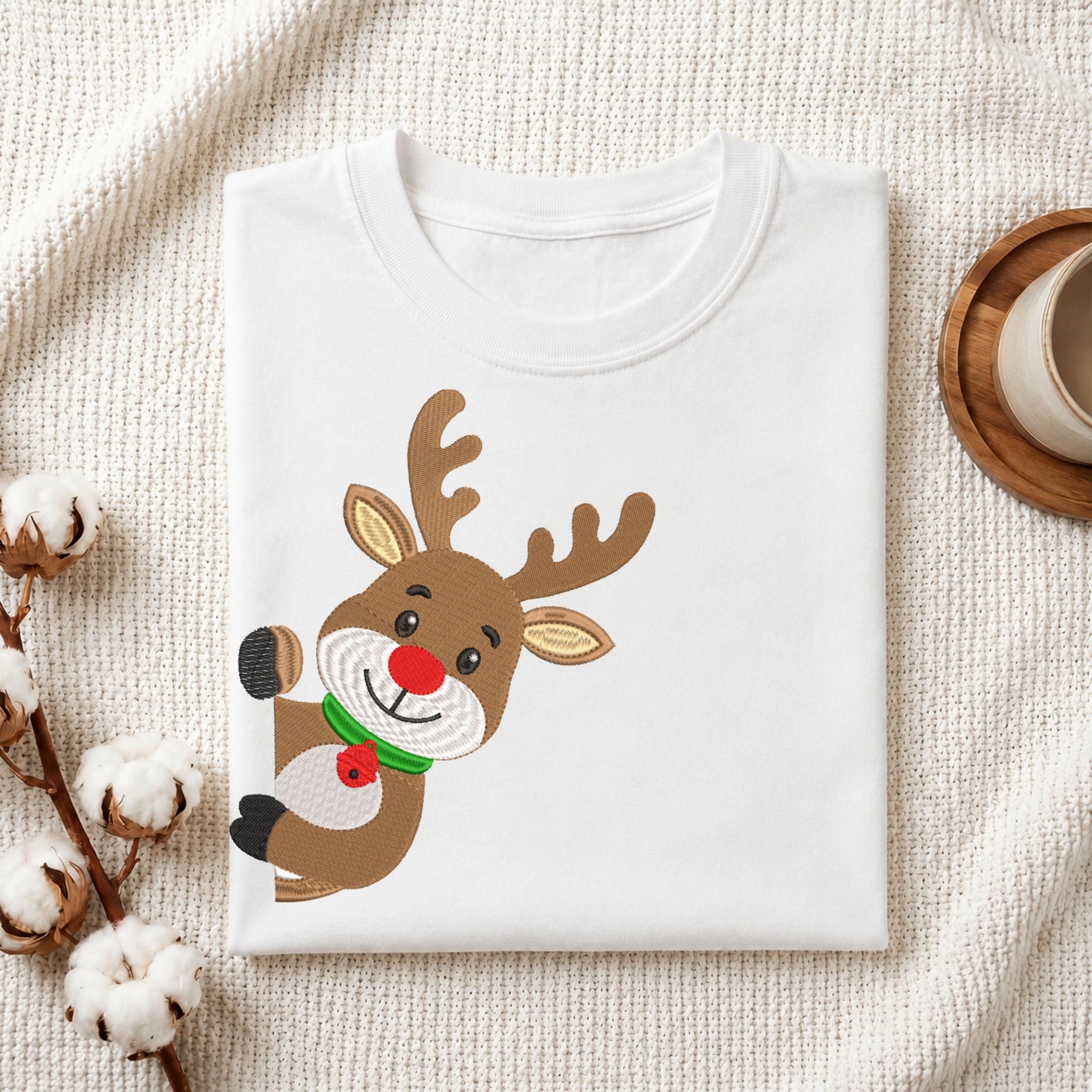 A folded white crewneck t-shirt with a cartoon reindeer graphic, styled on a textured cream fabric with cotton bolls and a wooden cup in the background.