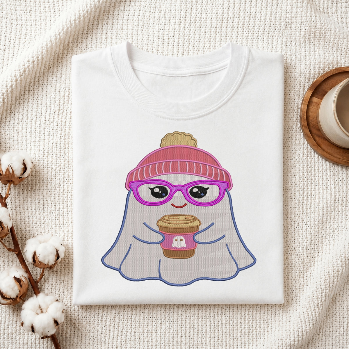 A folded white t-shirt with a cute cartoon ghost graphic wearing a pink beanie and purple glasses while holding a coffee cup, styled on a textured cream blanket with cotton bolls and a mug.