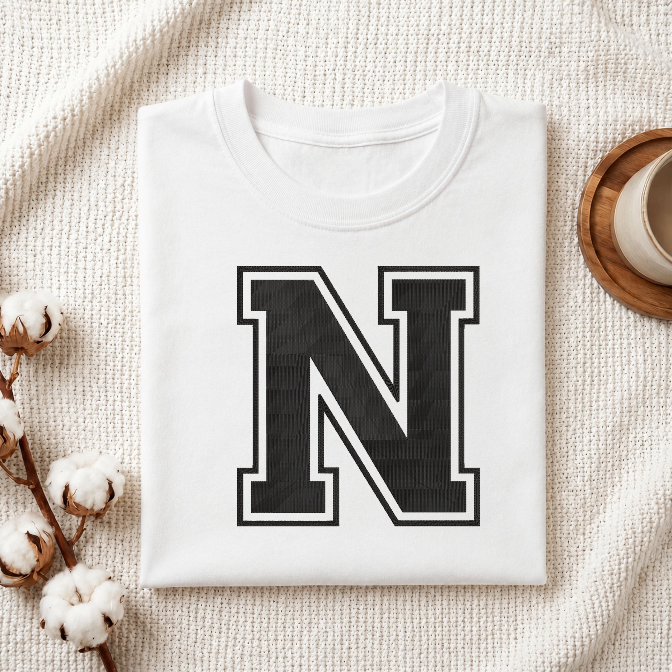 A folded white crewneck t-shirt with a large black block letter 'N' outlined in white on the front, styled on a textured cream knit blanket with cotton bolls and a cup on a wooden tray.