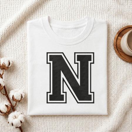 A folded white crewneck t-shirt with a large black block letter 'N' outlined in white on the front, styled on a textured cream knit blanket with cotton bolls and a cup on a wooden tray.