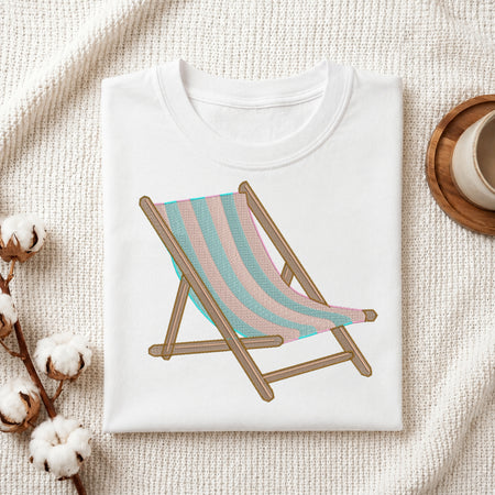 A folded white T-shirt laid flat on a textured cream fabric background, featuring a printed illustration of a wooden deck chair with pastel pink and aqua striped fabric. Cotton bolls and part of a wooden tray with a cup are visible as styled props.