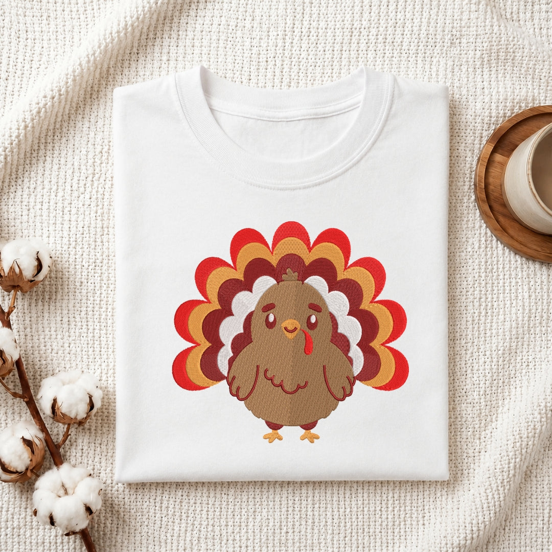 A folded white crewneck t-shirt featuring a cartoon turkey graphic with a red, orange, and brown feather tail, styled on a textured cream fabric with cotton bolls and a coffee cup.
