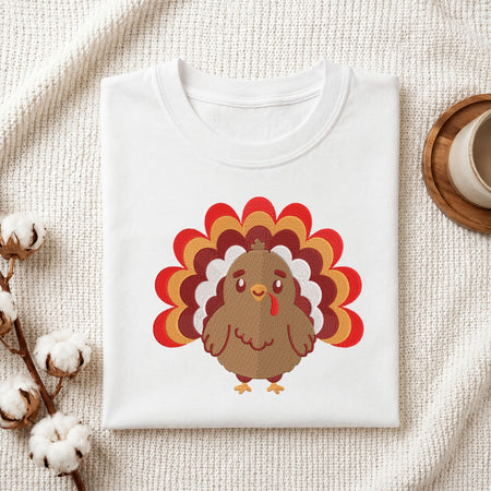 A folded white crewneck t-shirt featuring a cartoon turkey graphic with a red, orange, and brown feather tail, styled on a textured cream fabric with cotton bolls and a coffee cup.