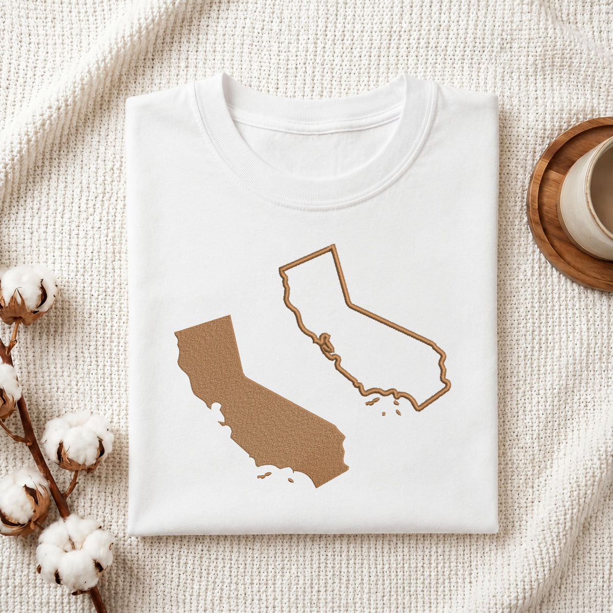 A folded white T-shirt laid flat on a textured cream fabric background, featuring a tan outline and filled silhouette of California on the front. Cotton bolls and part of a wooden cup are visible as styled props.