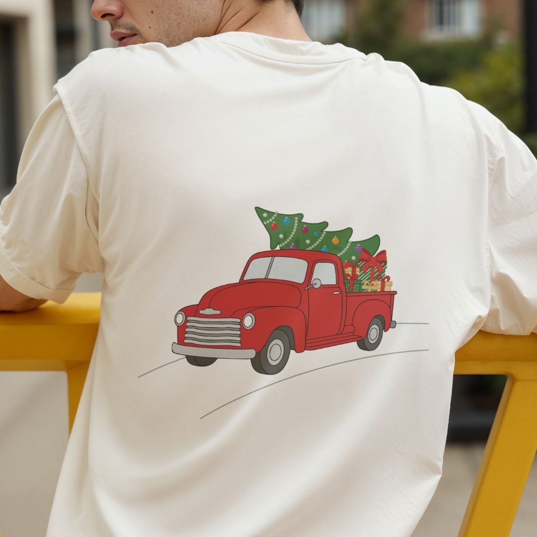 A man wearing a cream-colored t-shirt from the back, featuring a graphic of a red vintage truck carrying a green Christmas tree and holiday gifts.