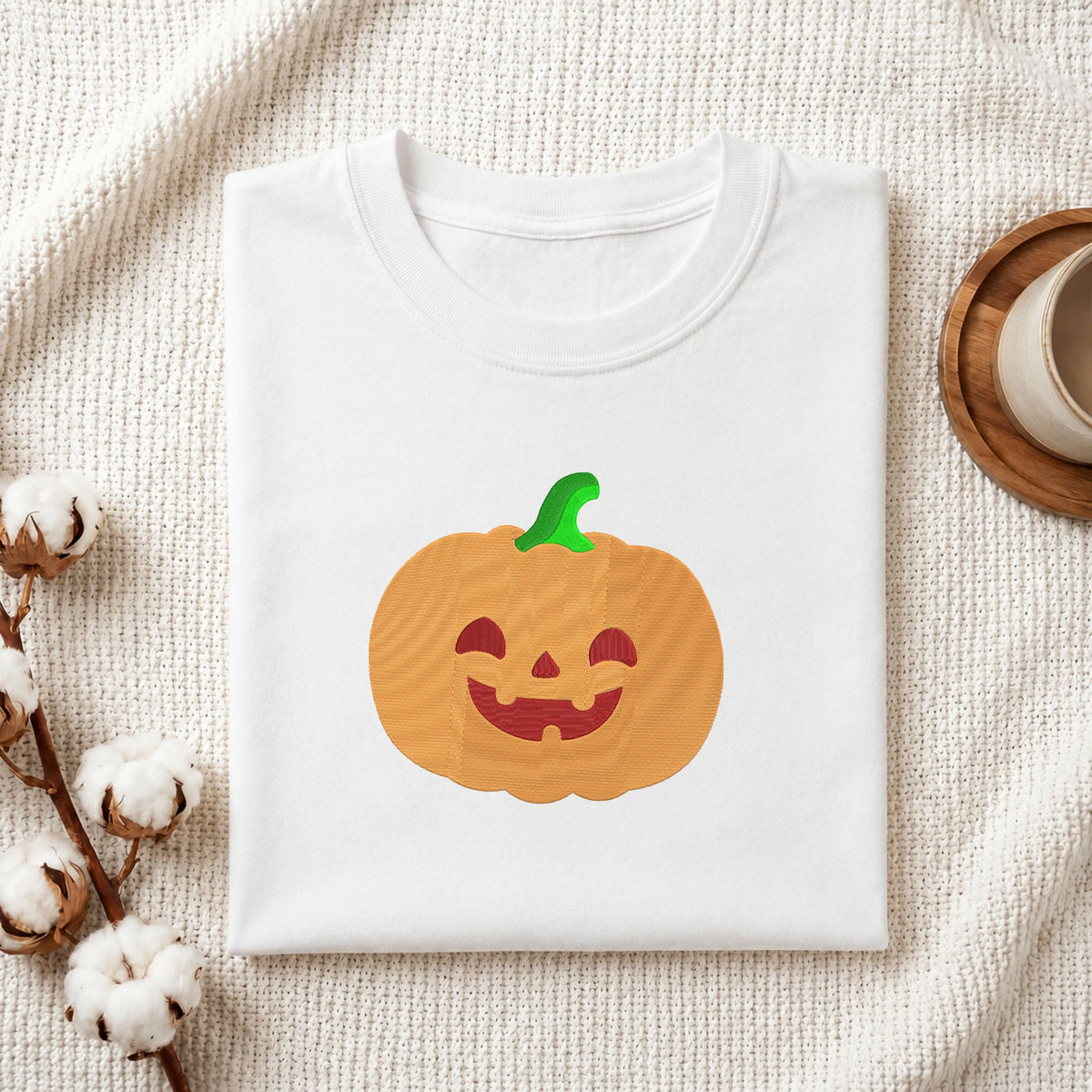 Folded white cotton t-shirt mockup featuring a cute jack-o'-lantern pumpkin machine embroidery design, styled with cotton stems and a wooden cup.   