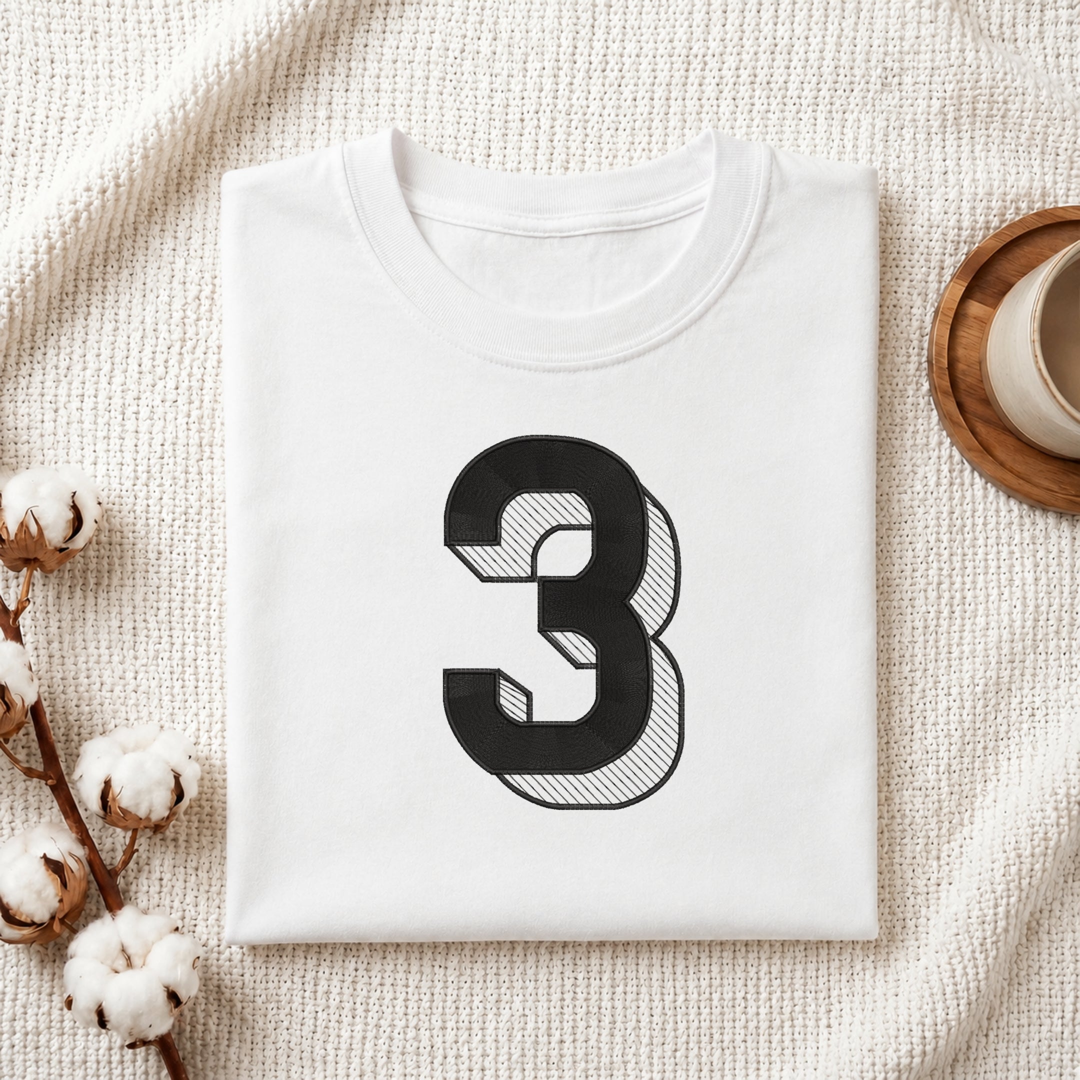 A neatly folded white crewneck t-shirt featuring a stitched 3D block number "3" design, styled with cotton branches and a wooden coaster.