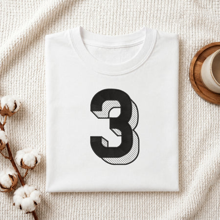 A neatly folded white crewneck t-shirt featuring a stitched 3D block number "3" design, styled with cotton branches and a wooden coaster.