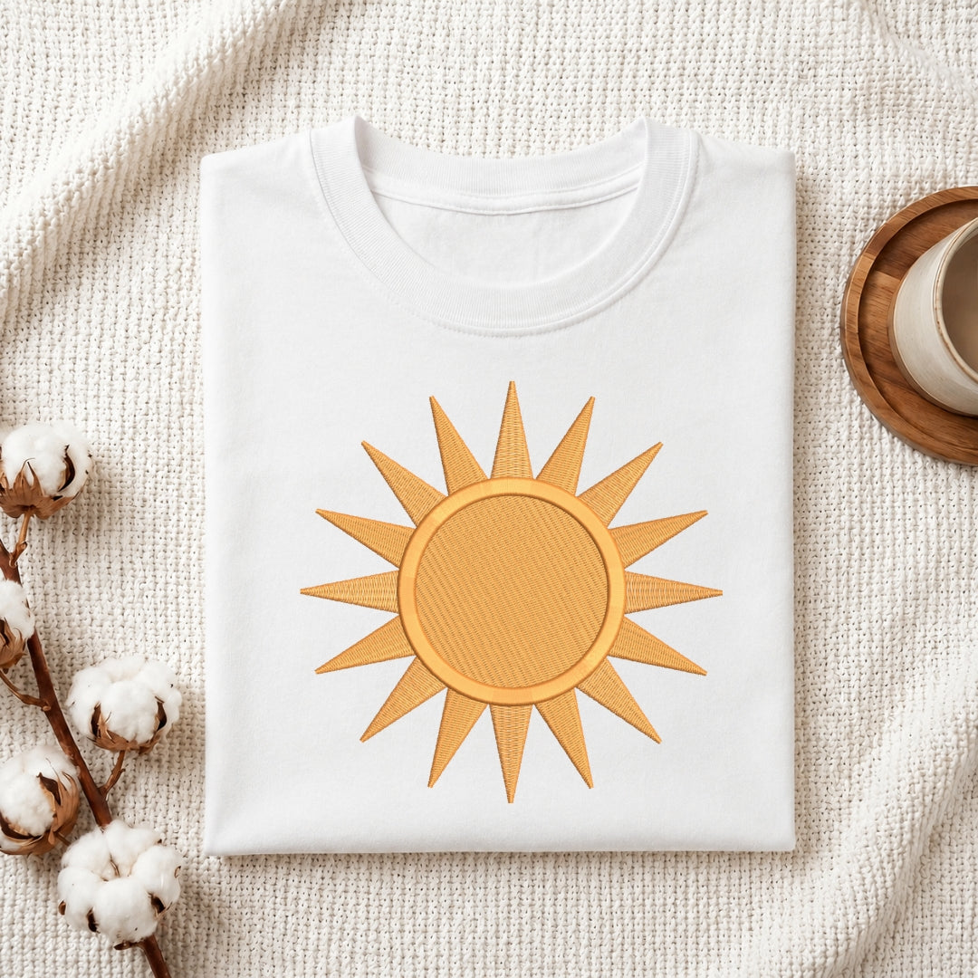 A folded white crewneck t-shirt featuring a golden-yellow embroidered sun design on the front, styled on a textured cream fabric background with cotton bolls and a wooden cup.