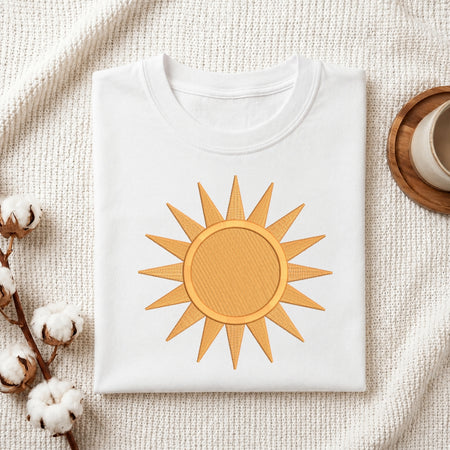 A folded white crewneck t-shirt featuring a golden-yellow embroidered sun design on the front, styled on a textured cream fabric background with cotton bolls and a wooden cup.