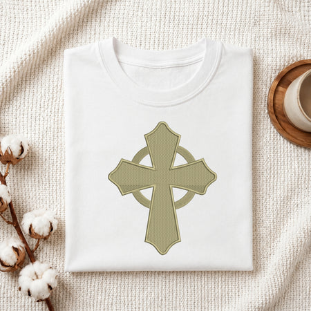  A neatly folded white crewneck t-shirt featuring the sage green Celtic cross embroidery design centered on the front, styled on a textured knit blanket next to a cup and cotton stem