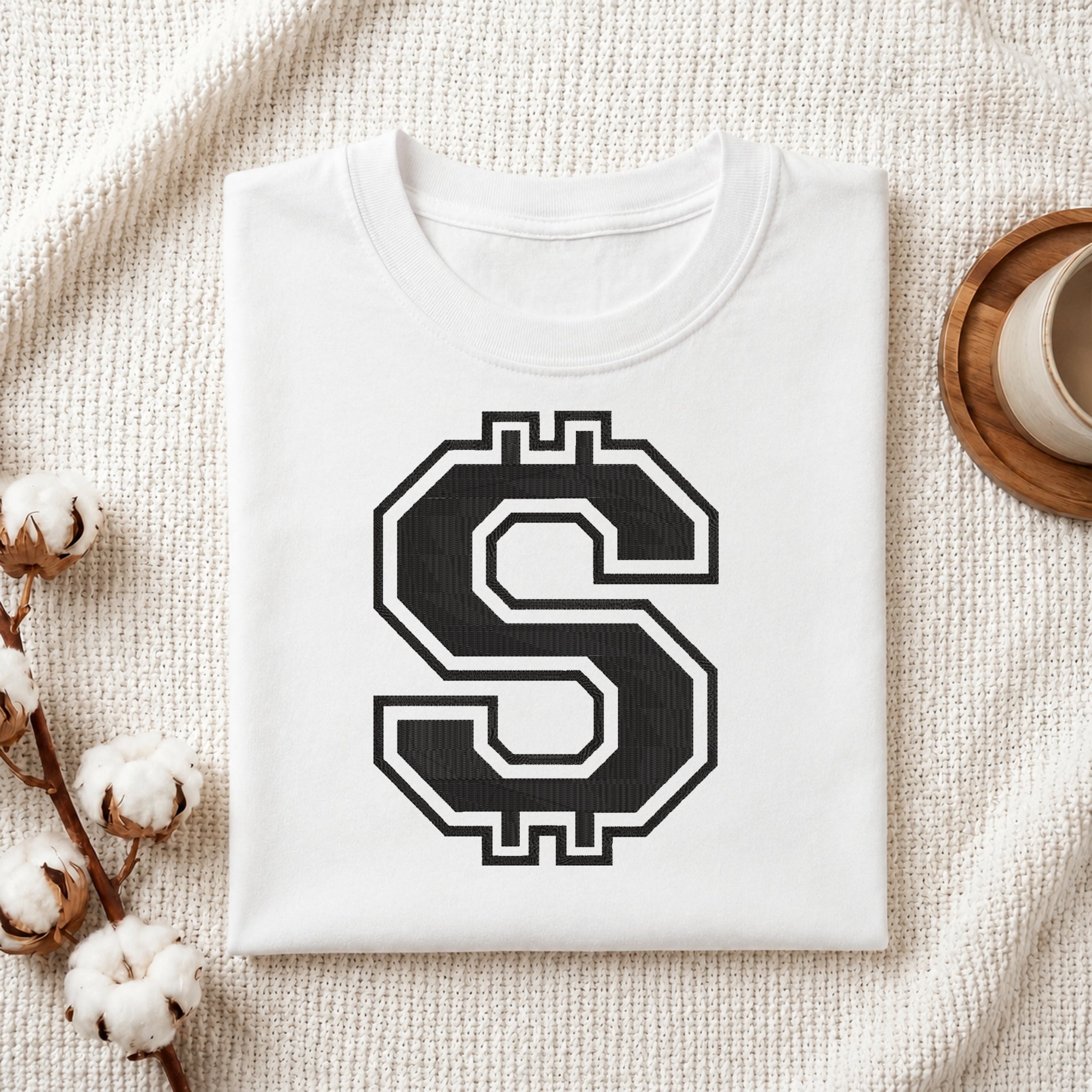 A folded white t-shirt with a large black and white stylized dollar sign graphic on the front, displayed on a textured cream knit blanket with decorative cotton bolls and a cup in the background.