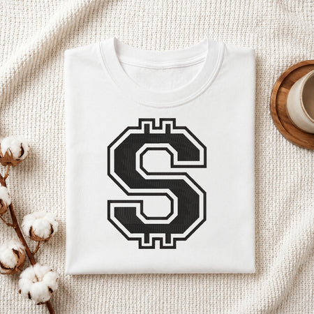 A folded white t-shirt with a large black and white stylized dollar sign graphic on the front, displayed on a textured cream knit blanket with decorative cotton bolls and a cup in the background.