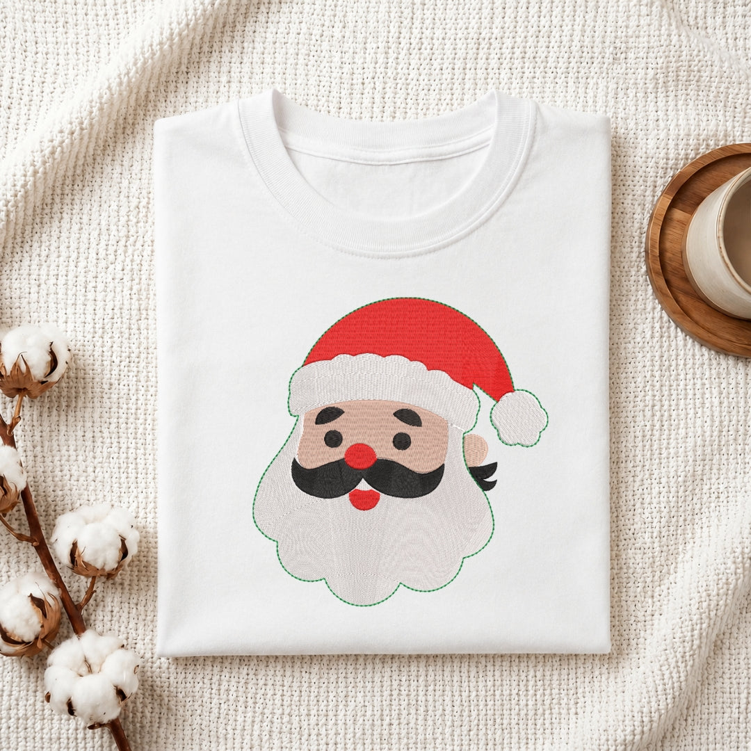 A folded white crewneck t-shirt featuring a cartoon Santa Claus face graphic on the front, styled in a flat-lay arrangement with cotton bolls and a cup on a textured cream fabric background.