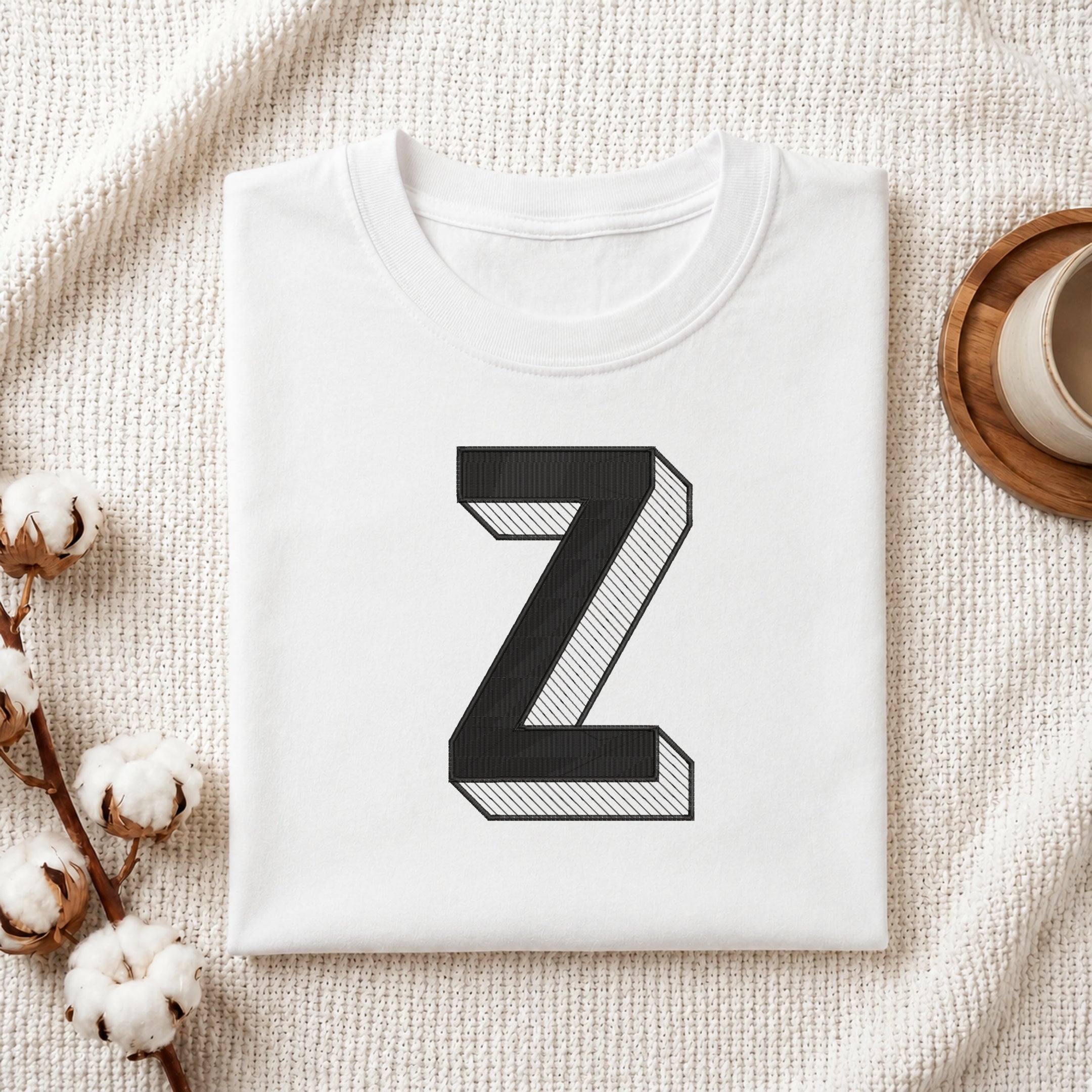 A folded white t-shirt with a large black block letter 'Z' printed on the front, styled on a textured cream fabric background with cotton bolls and a cup of coffee.