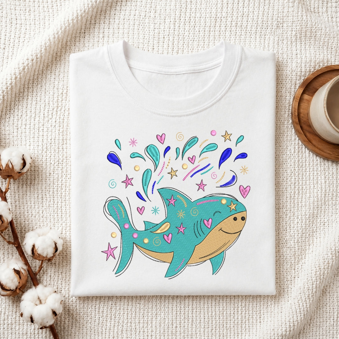A folded white crewneck t-shirt with a colorful cartoon shark graphic, styled on a textured cream fabric with cotton stems and a wooden cup in the background.