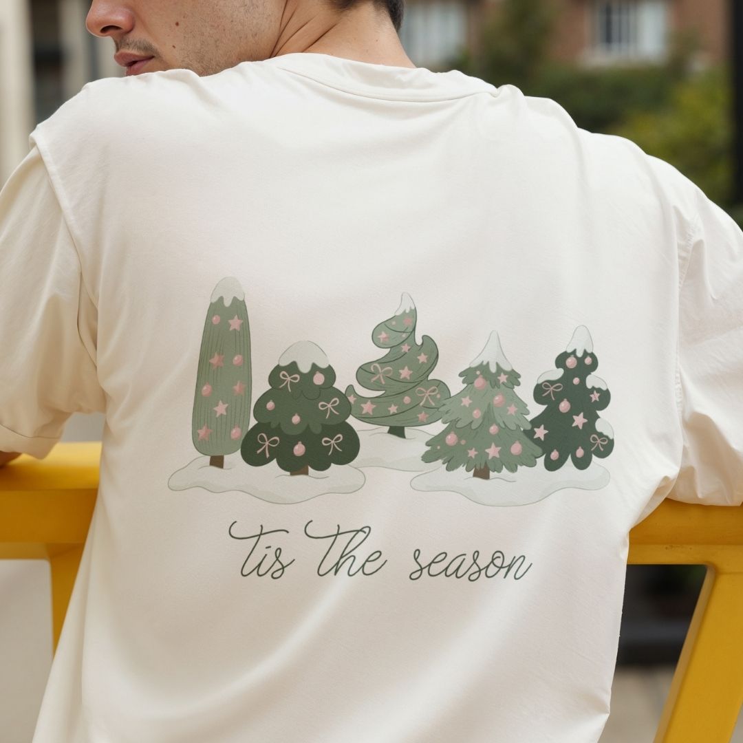 A man wearing a cream-colored t-shirt, showing the back of the shirt with a graphic of four decorated Christmas trees in shades of green and pink, above the cursive text 'Tis the season'.
