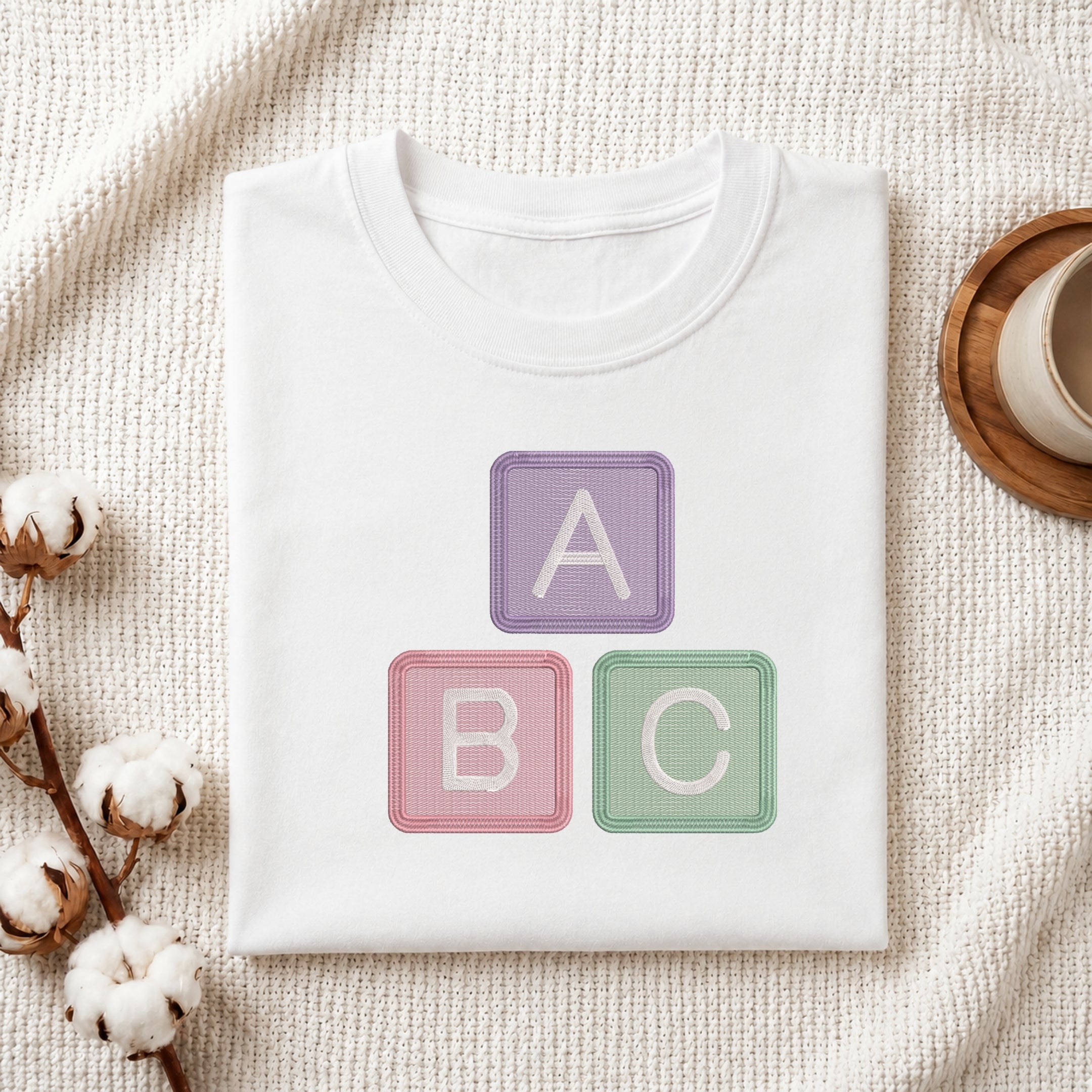 A folded white crewneck t-shirt with a graphic of three pastel-colored baby blocks reading 'A', 'B', and 'C' in purple, pink, and green. The t-shirt is styled in a flat-lay on a textured cream knit blanket with cotton bolls and a wooden cup on a coaster.