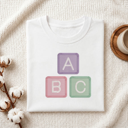 A folded white crewneck t-shirt with a graphic of three pastel-colored baby blocks reading 'A', 'B', and 'C' in purple, pink, and green. The t-shirt is styled in a flat-lay on a textured cream knit blanket with cotton bolls and a wooden cup on a coaster.