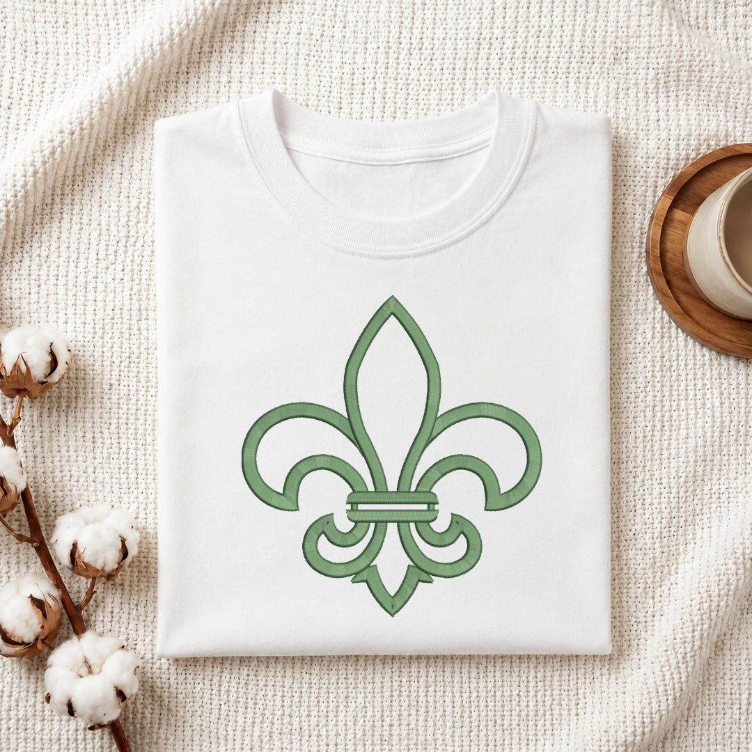 A folded white crewneck t-shirt with a green fleur-de-lis graphic on the front, styled on a textured cream knit blanket with cotton bolls and a wooden cup in the background.