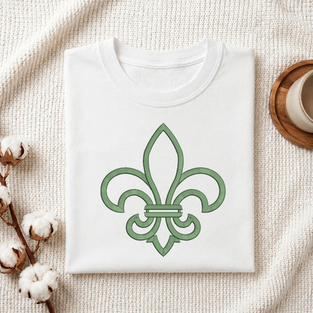 A folded white crewneck t-shirt with a green fleur-de-lis graphic on the front, styled on a textured cream knit blanket with cotton bolls and a wooden cup in the background.