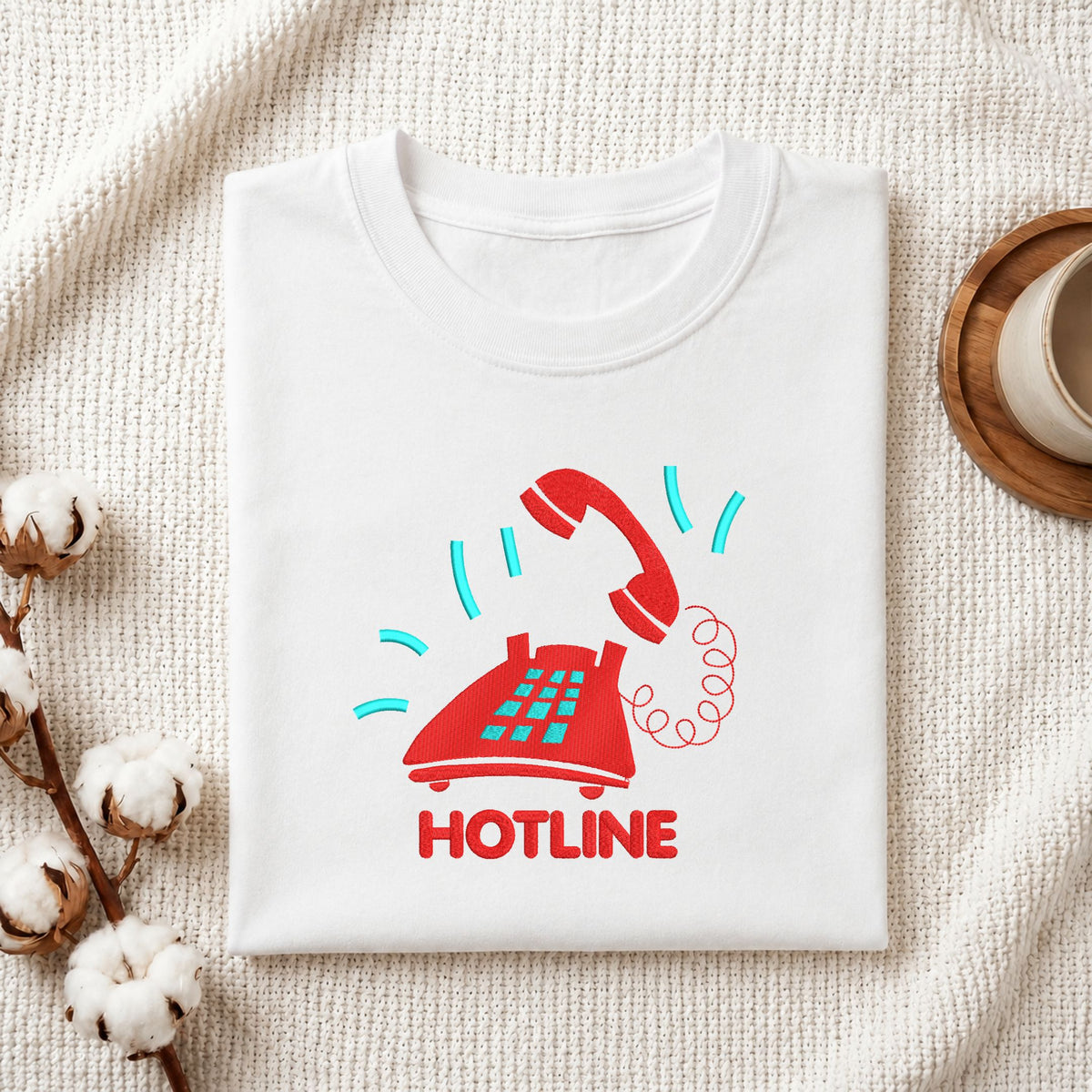  Folded white cotton t-shirt mockup featuring a red vintage telephone and "HOTLINE" machine embroidery design, styled with cotton stems and a wooden cup   