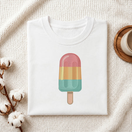A folded white T-shirt laid flat on a textured cream fabric background, featuring a colorful popsicle graphic on the front. Cotton bolls and part of a wooden cup are visible as styled props around the shirt.