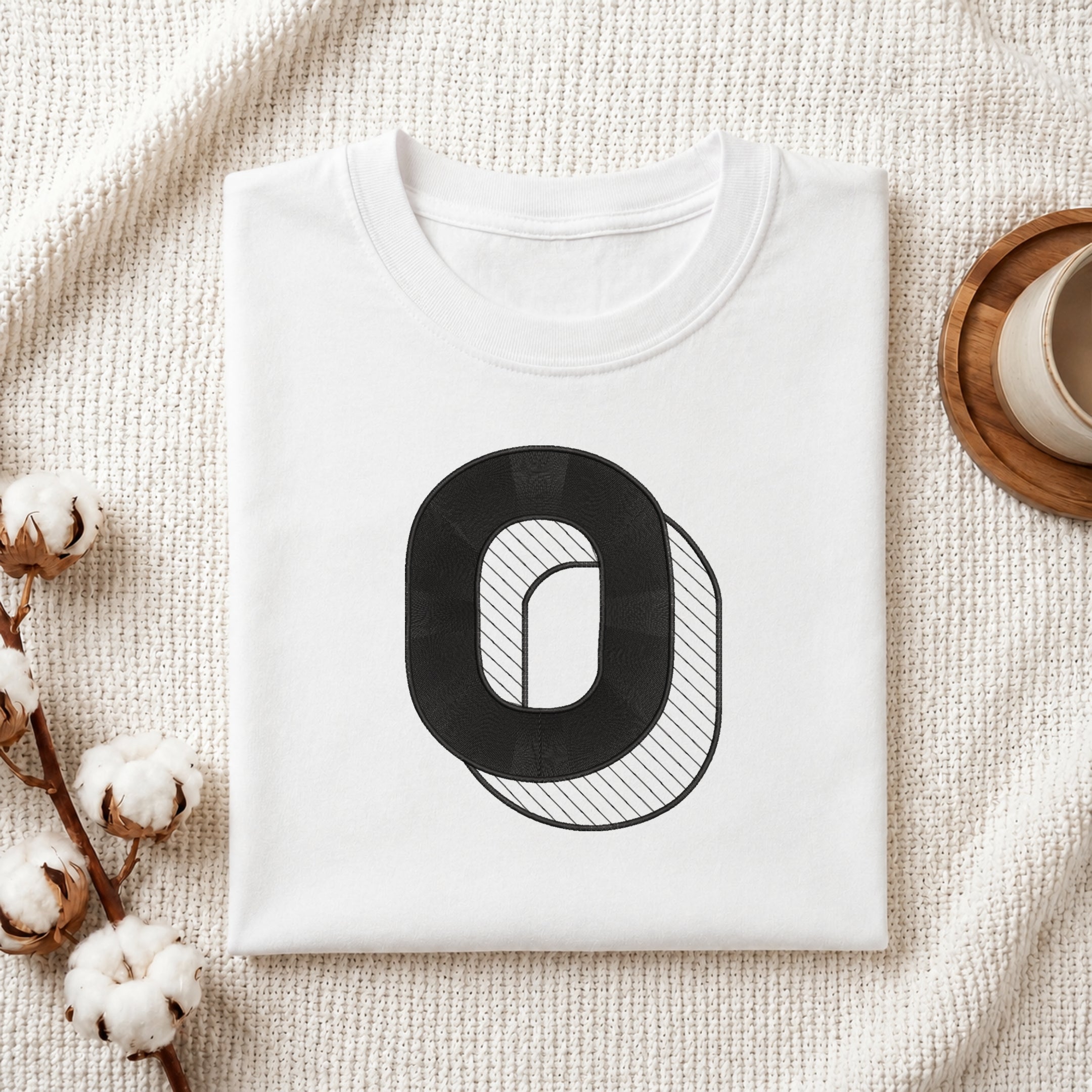 A folded white crewneck t-shirt with a black graphic letter 'O' on the front, styled on a textured cream knit blanket with cotton bolls and a wooden cup in the background.