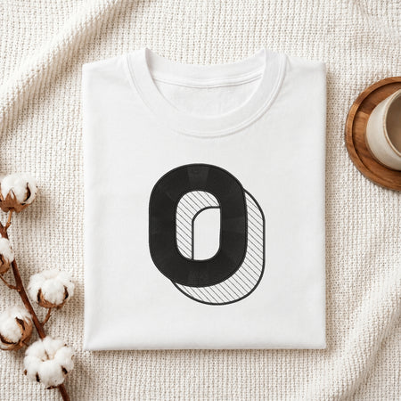 A folded white crewneck t-shirt with a black graphic letter 'O' on the front, styled on a textured cream knit blanket with cotton bolls and a wooden cup in the background.