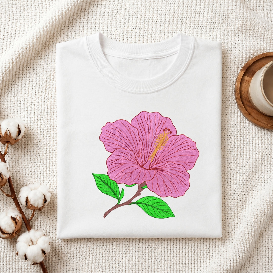 A folded white crewneck t-shirt featuring a graphic of a pink hibiscus flower with green leaves, styled on a textured cream fabric with cotton bolls and a wooden cup in the background.