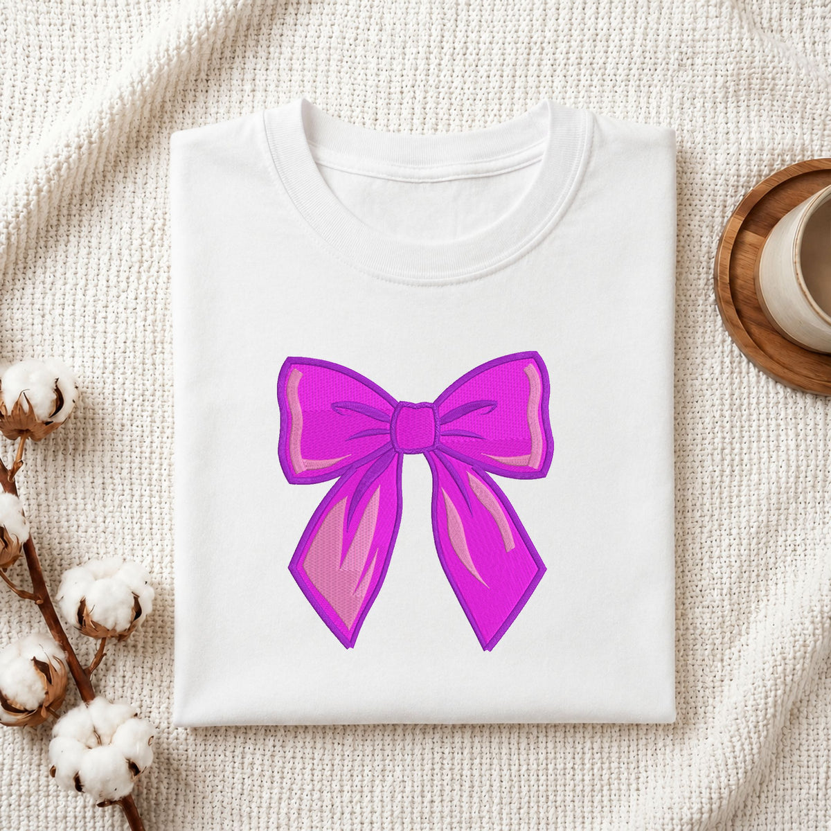 Folded white cotton t-shirt mockup featuring a vibrant purple bow machine embroidery design, styled with cotton stems and a wooden cup.