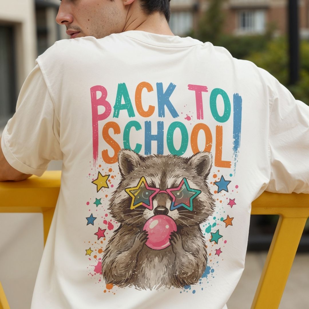 A man wearing a cream-colored t-shirt from the back, featuring a colorful graphic print that reads 'BACK TO SCHOOL' above an illustration of a raccoon wearing star-shaped sunglasses and blowing pink bubblegum. The photo is taken outdoors in a sunny, blurred environment.
