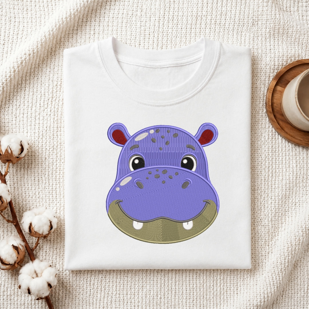 A folded white crewneck t-shirt featuring a cute cartoon hippo face graphic on the front, styled on a textured cream fabric background with cotton bolls and a wooden cup.