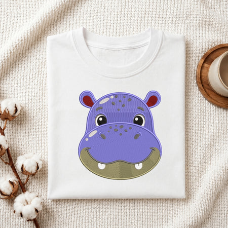 A folded white crewneck t-shirt featuring a cute cartoon hippo face graphic on the front, styled on a textured cream fabric background with cotton bolls and a wooden cup.